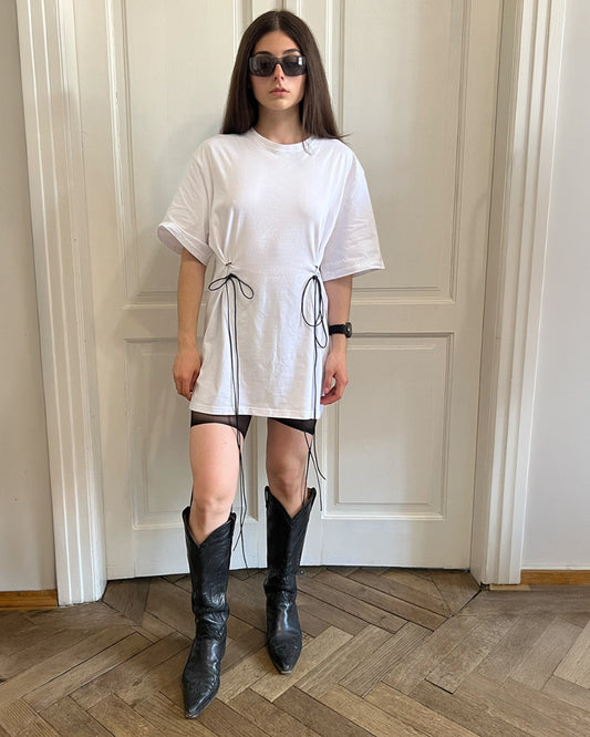 A woman stands indoors in front of a white paneled door wearing an oversized white T-shirt dress with black side drawstrings, black fitted shorts, black knee-high boots, and dark sunglasses.